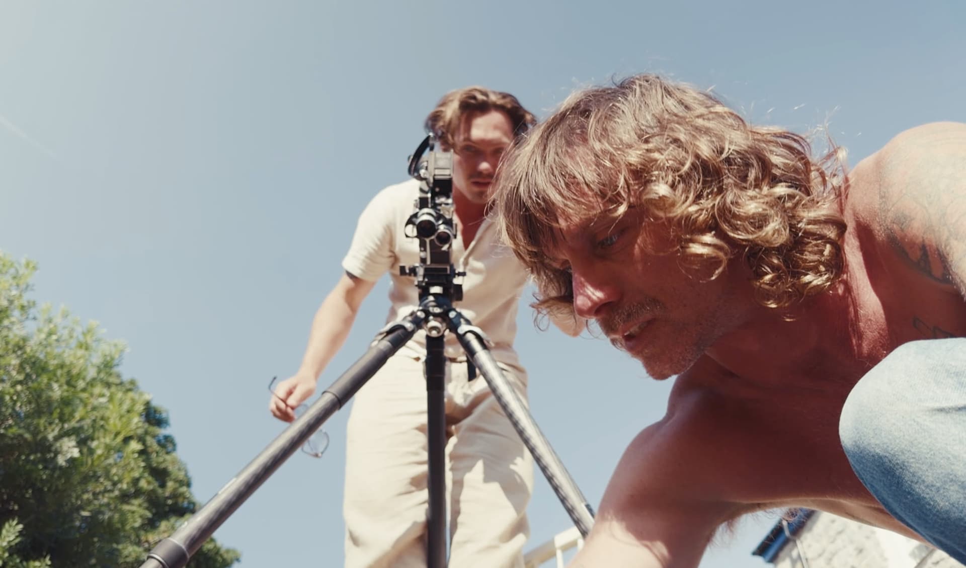 Two of the crew filming on location in bright sun, one behind a cine camera on a tripod and one crouched close to the lens