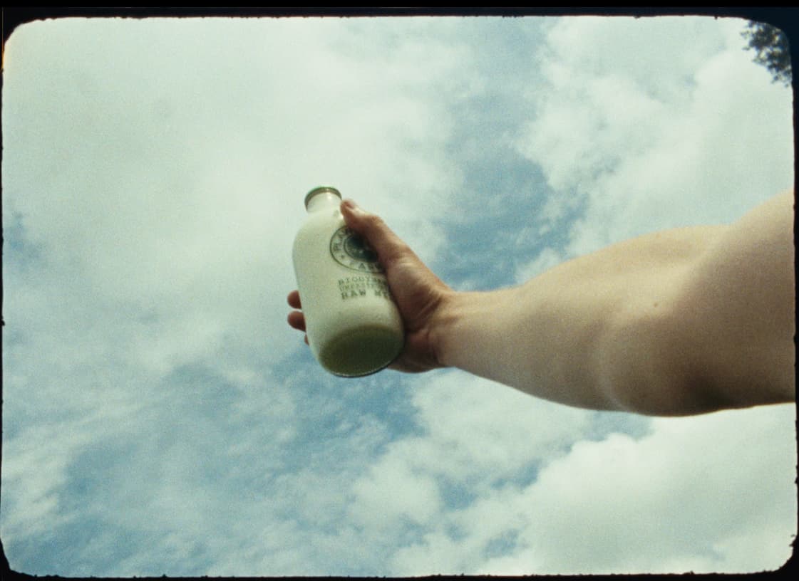 A hand holds a bottle of raw milk up against a bright, cloud-filled sky
