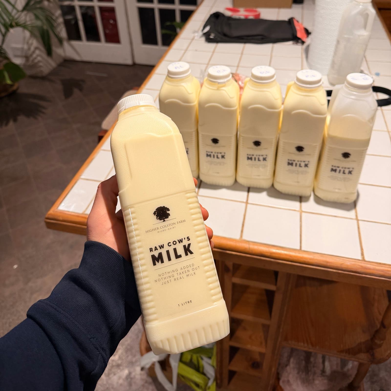 A hand holding a liter bottle of Higher Colston Farm raw cow’s milk, more bottles lined up behind