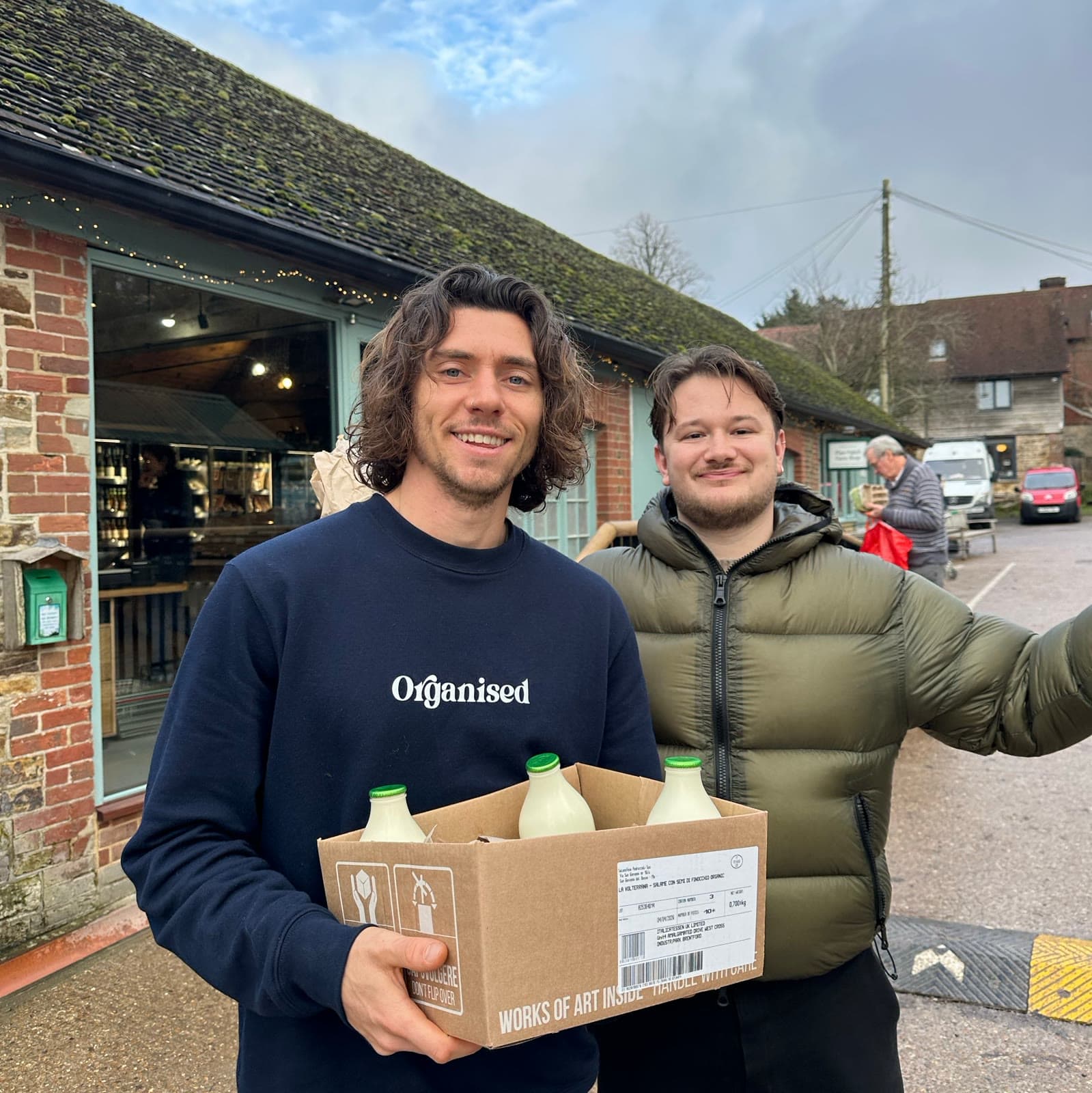 Two members smiling outside the Plaw Hatch farm shop with a box of raw milk bottles