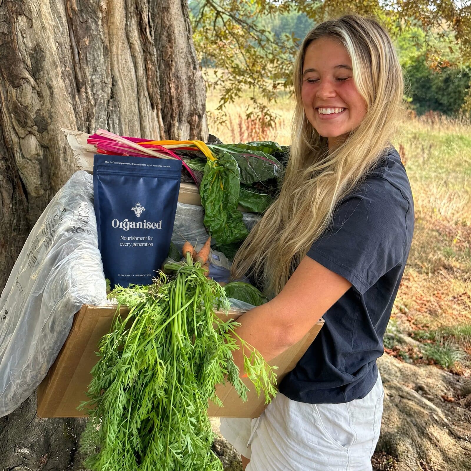 A smiling member holding a box of colorful seasonal veg beneath a tree at My Little Farm