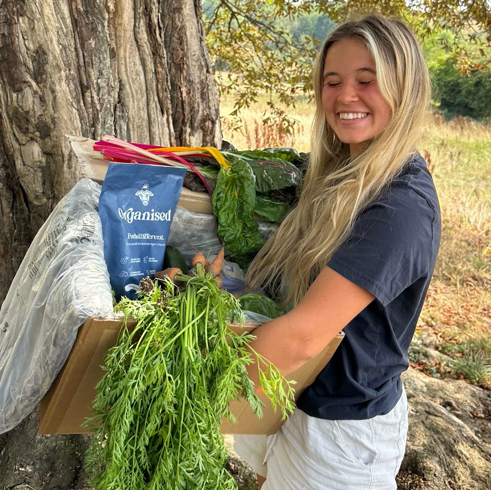 A smiling member holding a box of colourful seasonal veg beneath a tree at My Little Farm