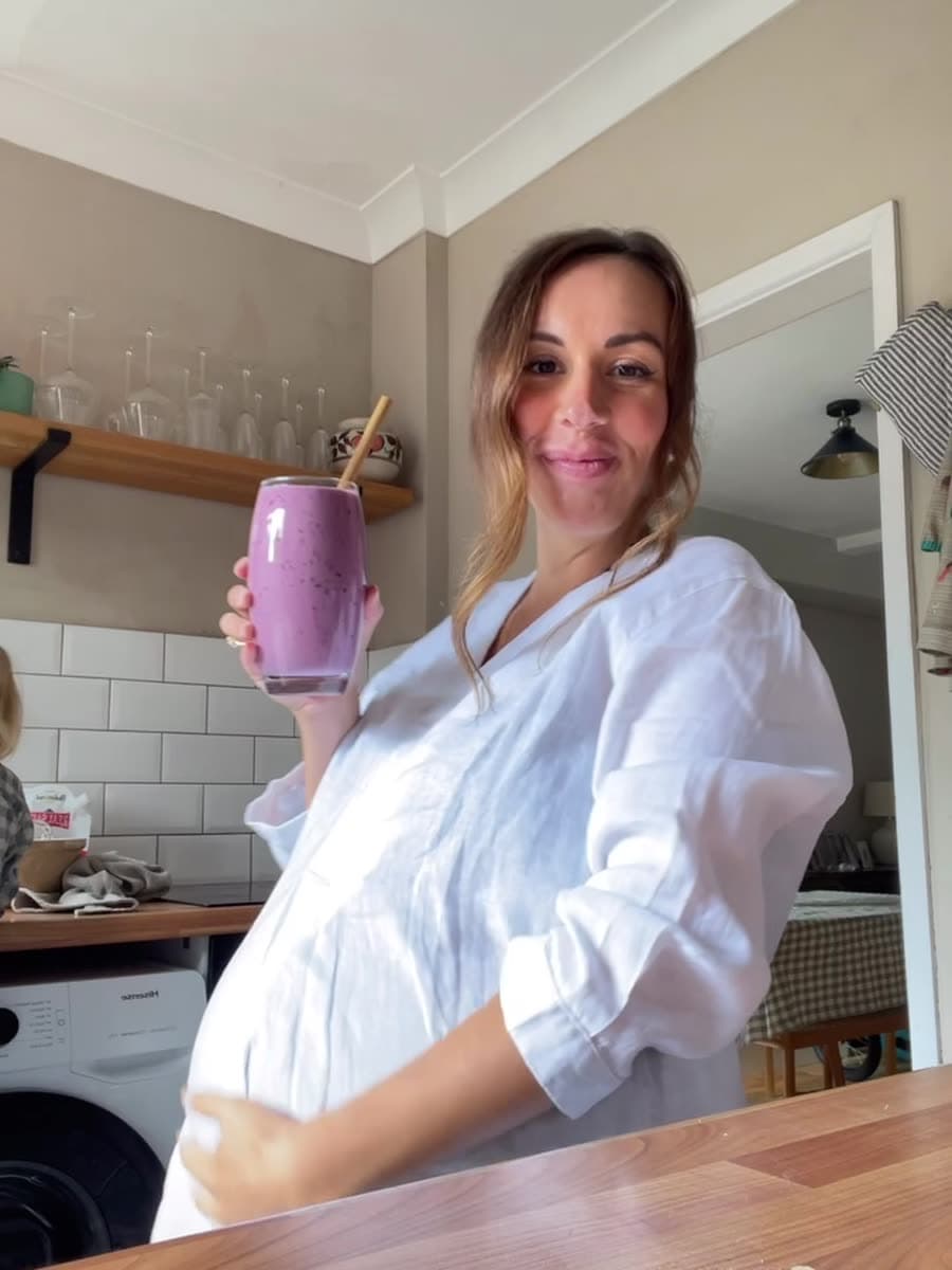 Jess, pregnant in a white linen shirt, smiling in her kitchen and holding up a berry smoothie