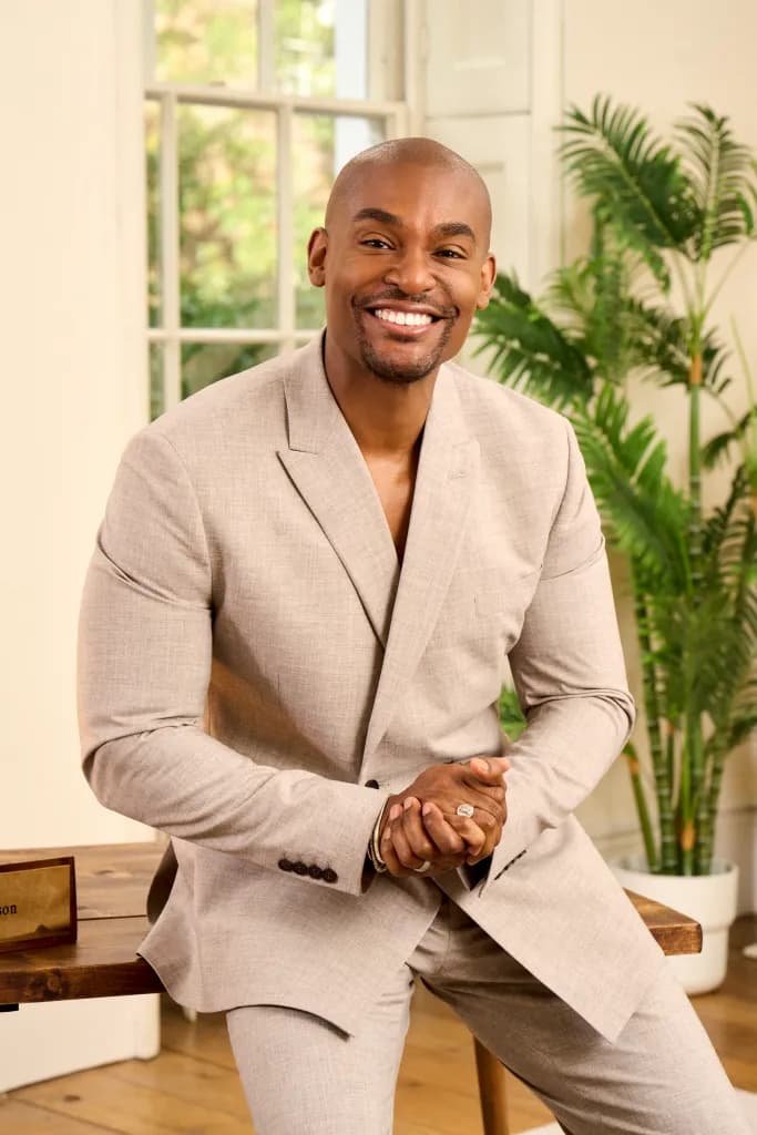 Paul Brunson in a pale suit, smiling, sitting on the edge of a wooden table beside a tall plant