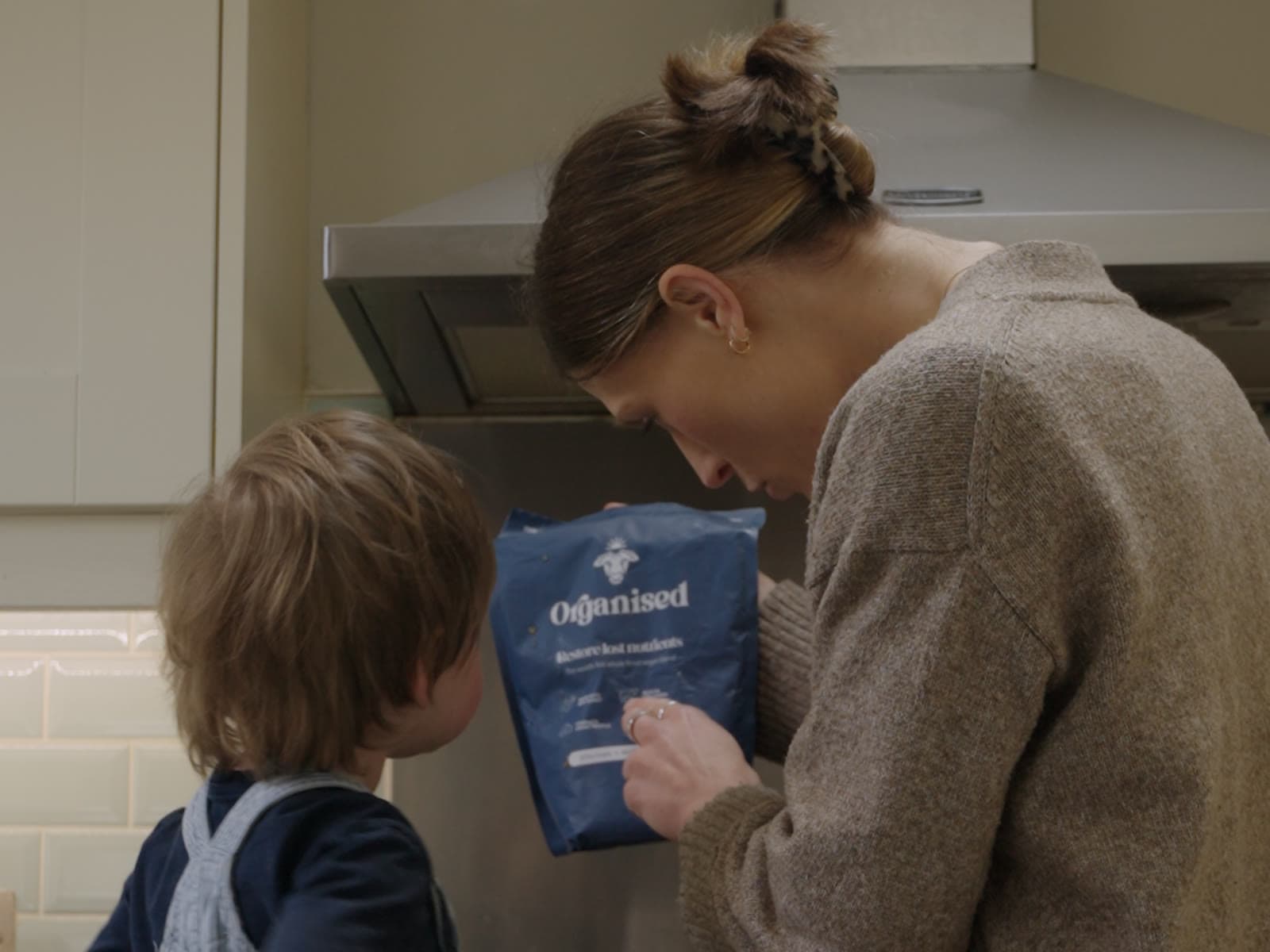 A mother at the stove showing an Organised pouch to her toddler, who watches from a kitchen step