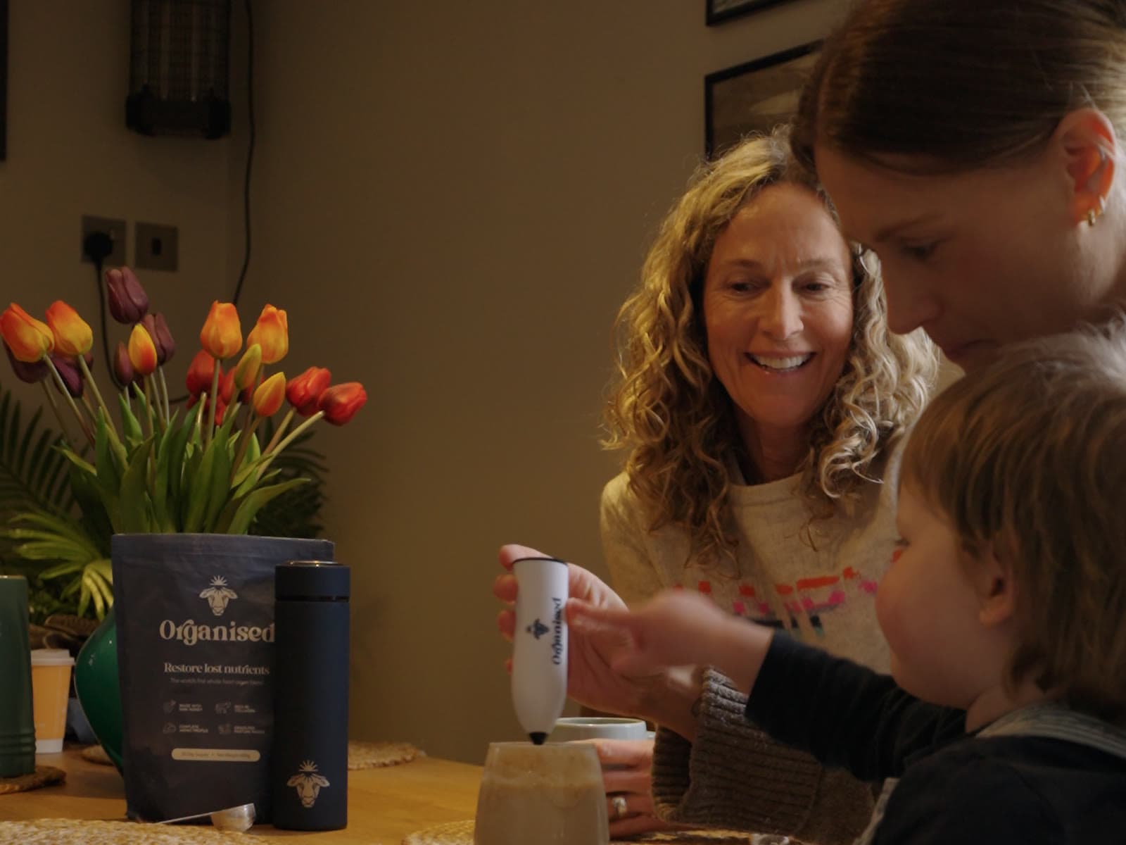A family at a kitchen table, a toddler on his mother's lap frothing a drink with the Organised whisk beside the pouch and bottle
