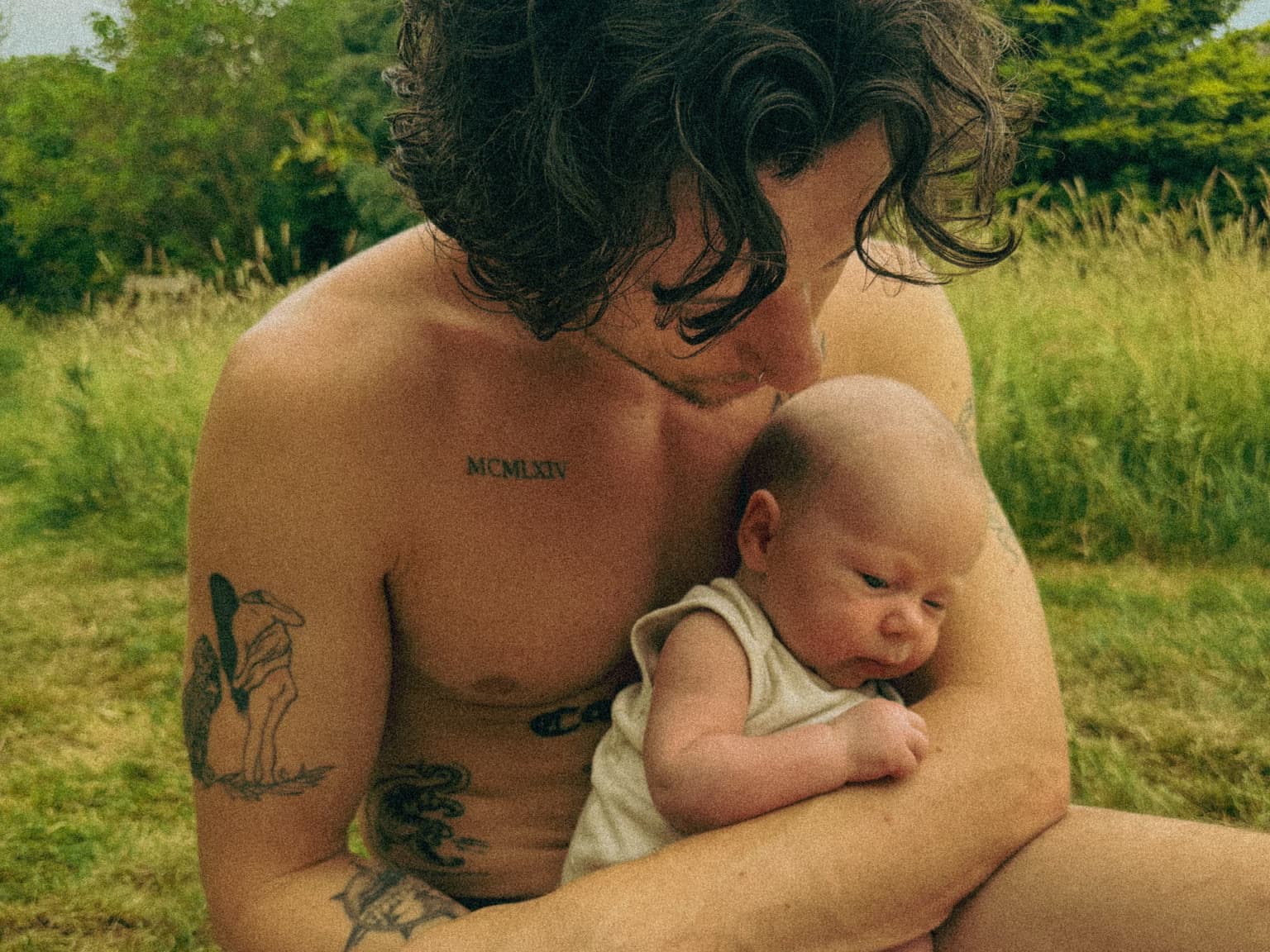 A father sitting on a blanket in a meadow, holding his baby against his chest