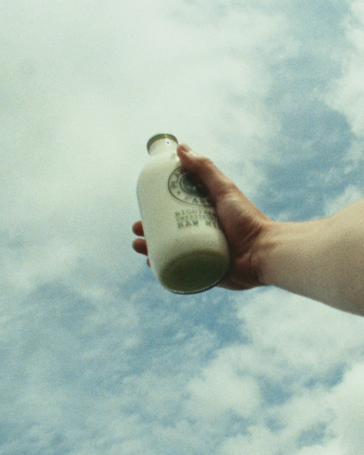 A hand holds a bottle of raw milk up against a bright, cloud filled sky
