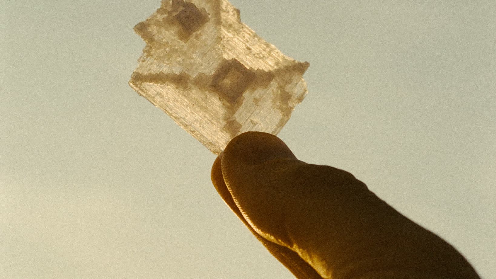 A single translucent salt crystal held between finger and thumb, backlit gold against a pale sky